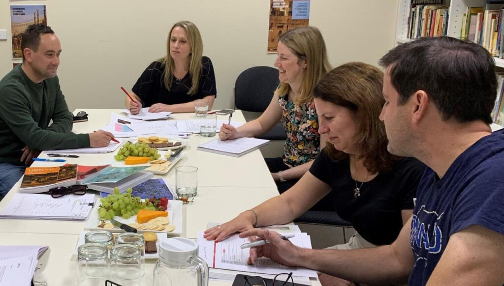Five people sit around a boardroom table. They have textbooks and documents in front of them. The Group are working on creating a new textbook together.